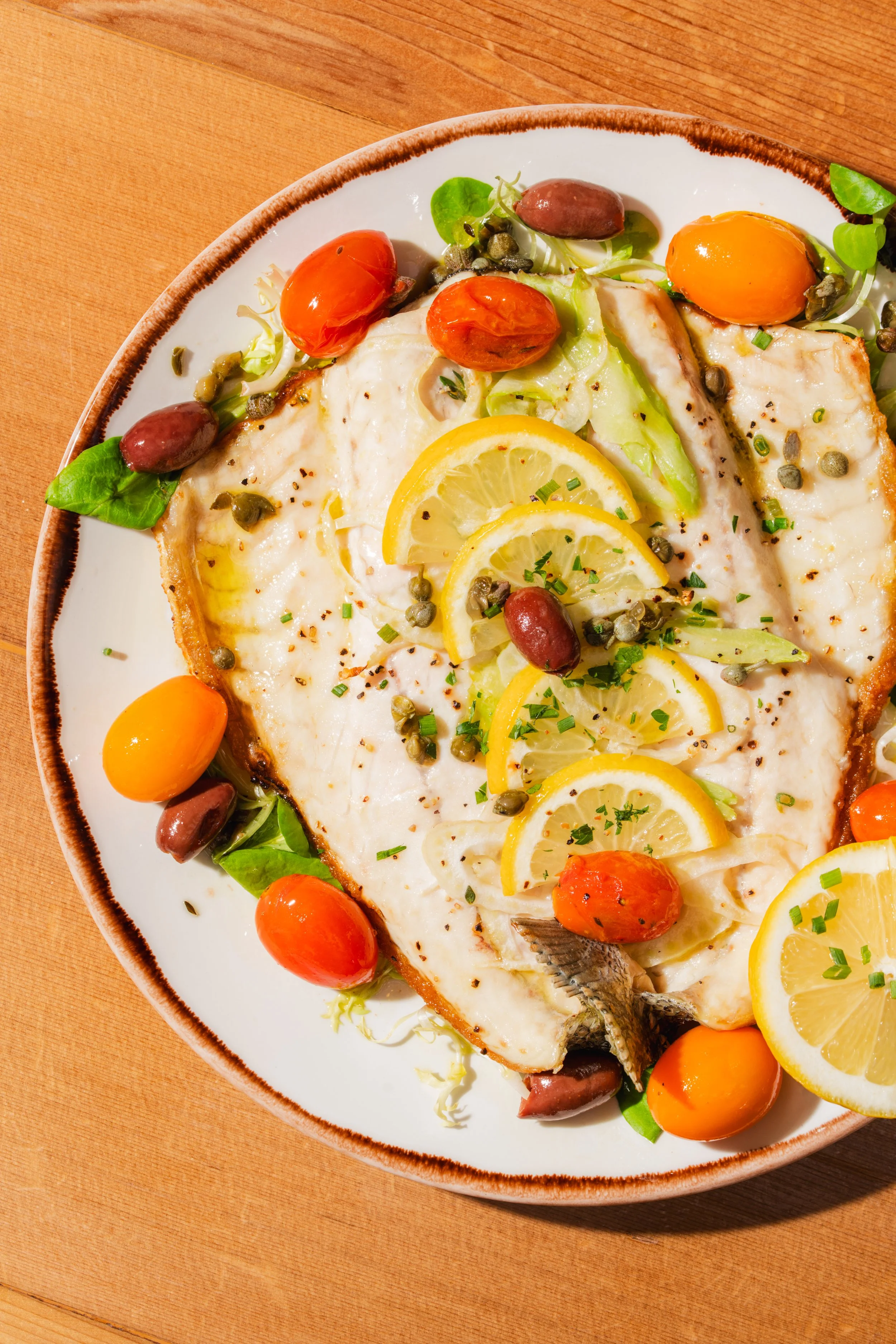 A fish fillet dish garnished with lemon slices, cherry tomatoes, olives, capers, and herbs on a white plate with a brown rim, placed on a wooden and orange table.
