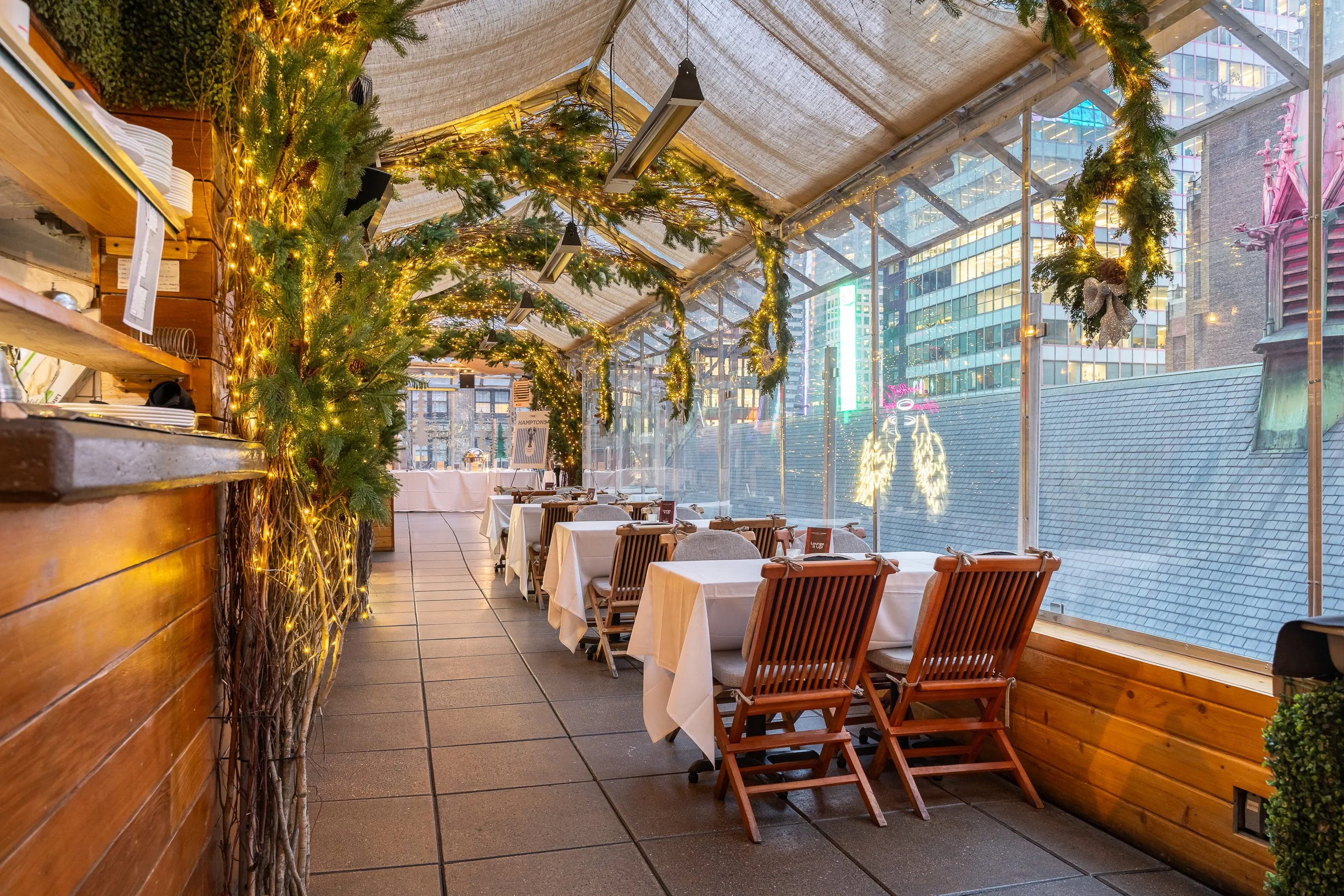 Empty restaurant with white tablecloths, wooden chairs, and holiday decorations including green garlands with lights and ornaments inside a glass-enclosed space of a city building.