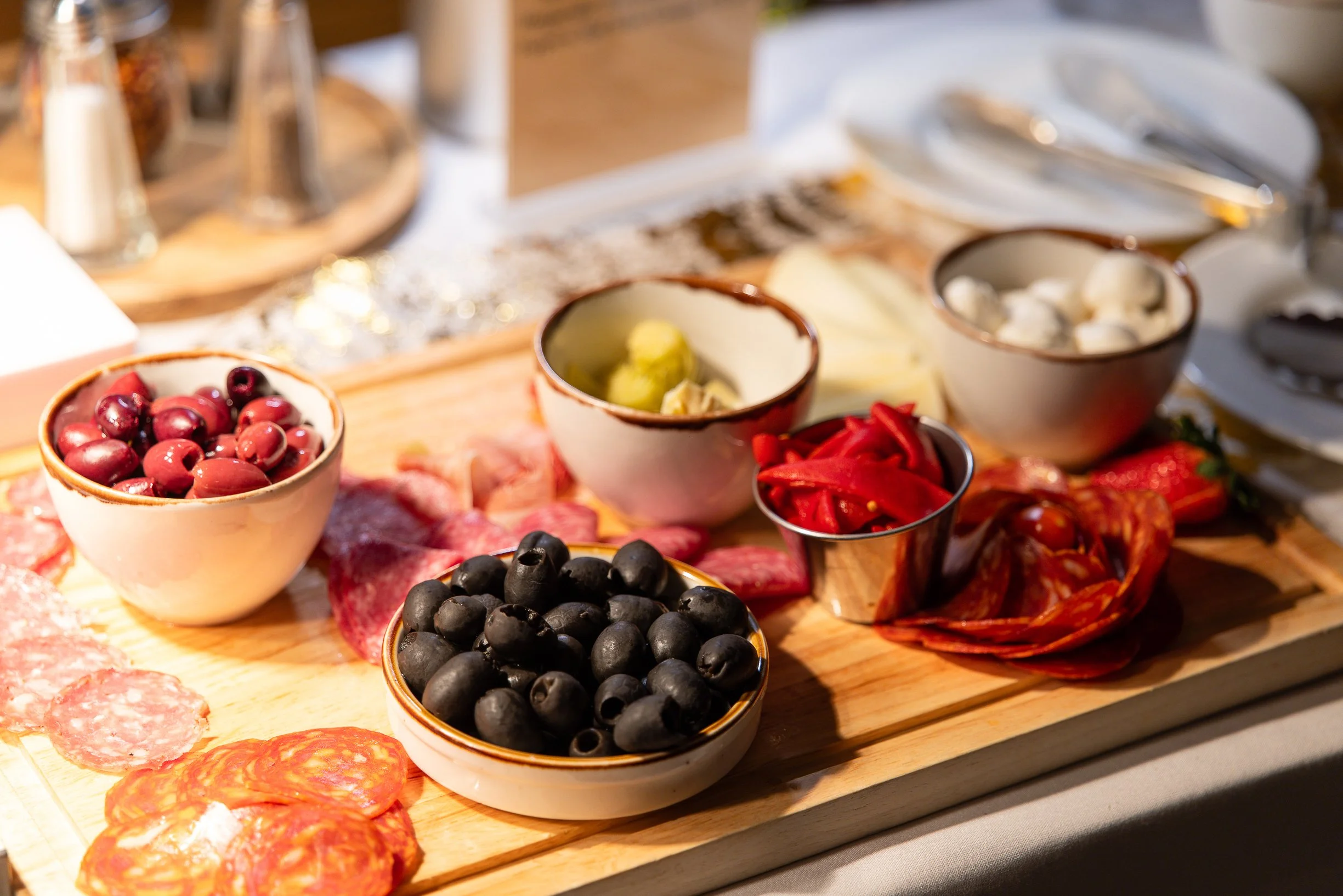 A wooden board with bowls of cherries, blueberries, and black olives, along with slices of cured meat and red peppers, part of a charcuterie spread.