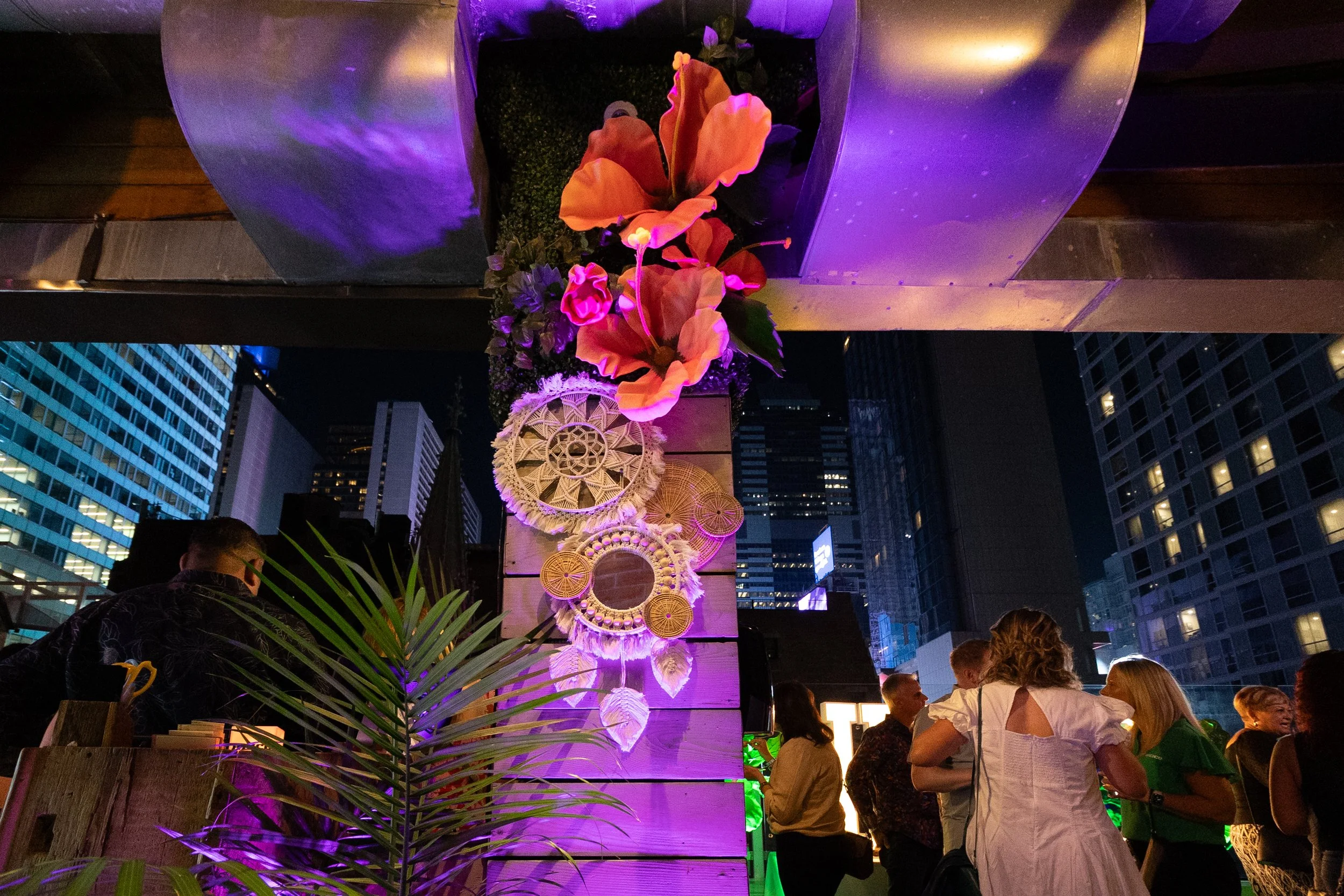 Night scene at an outdoor event with colorful lighting, featuring a vertical display of decorative flowers and woven circular decorations on a wooden pillar, surrounded by people in conversation in a city with tall buildings.