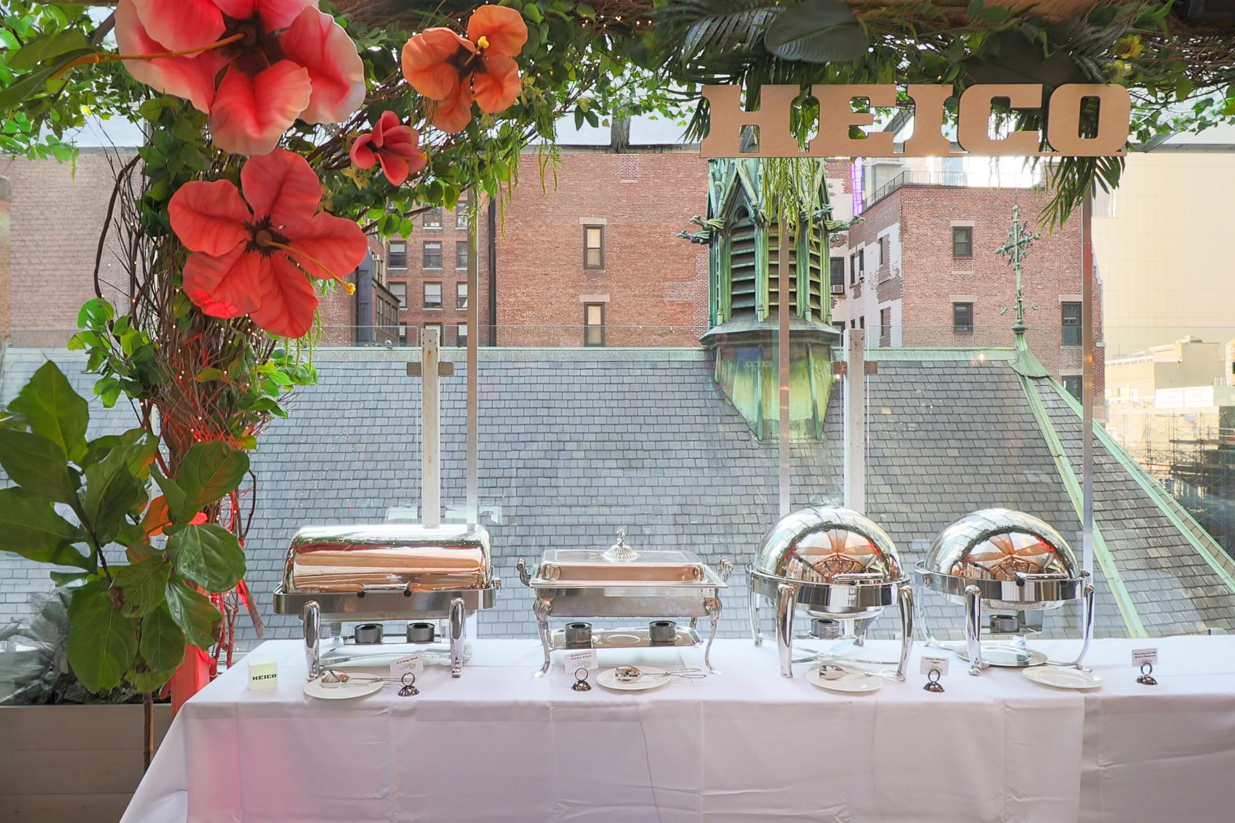 A buffet table with serving trays and platters, decorated with a large floral arrangement of pink and orange flowers and green leaves at the side, set against a city rooftop backdrop with buildings and a brick wall.