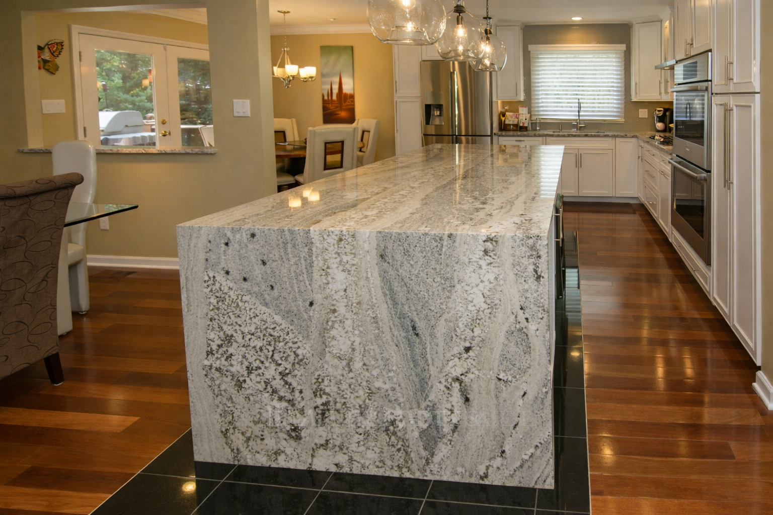Modern kitchen with a large granite island, white cabinets, stainless steel appliances, wooden flooring, and a window with blinds.
