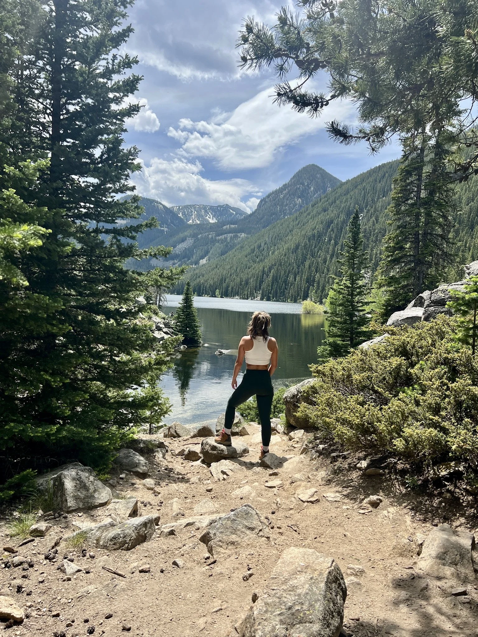 A woman standing on a rocky trail overlooking a lake surrounded by dense green pine trees and tall mountains under a partly cloudy sky.