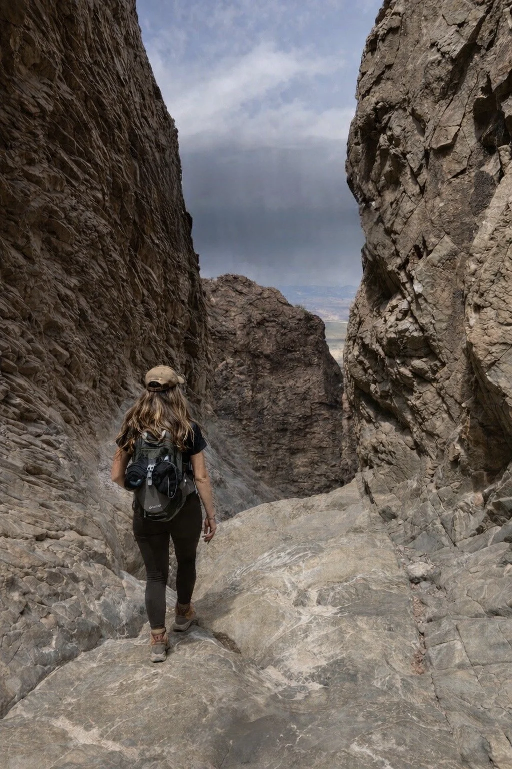 Woman hiking through a narrow rocky canyon with steep walls, wearing a hat, black shirt, jeans, carrying a backpack, and looking ahead as she walks on the rocky terrain.