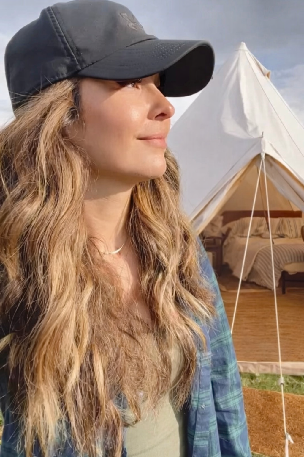 A woman with long, wavy, blonde hair and a black cap standing outdoors near a camping tent, with a bedroom visible inside the tent.