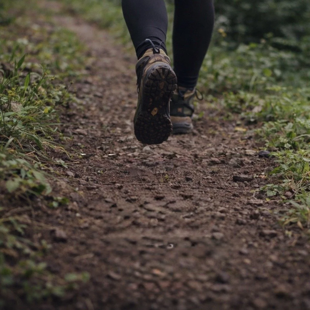 Close-up of a person running on a dirt trail in a wooded area, wearing hiking shoes and black leggings.