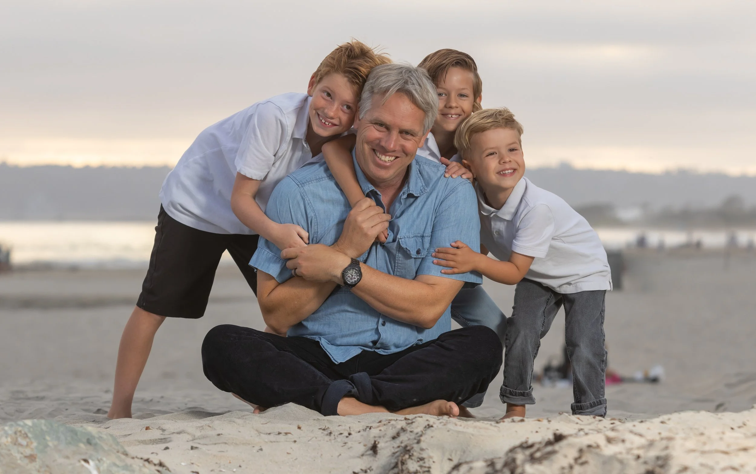 Smiling man sitting on the beach with three young boys, all smiling and leaning on him, with a cloudy sky and the ocean in the background.