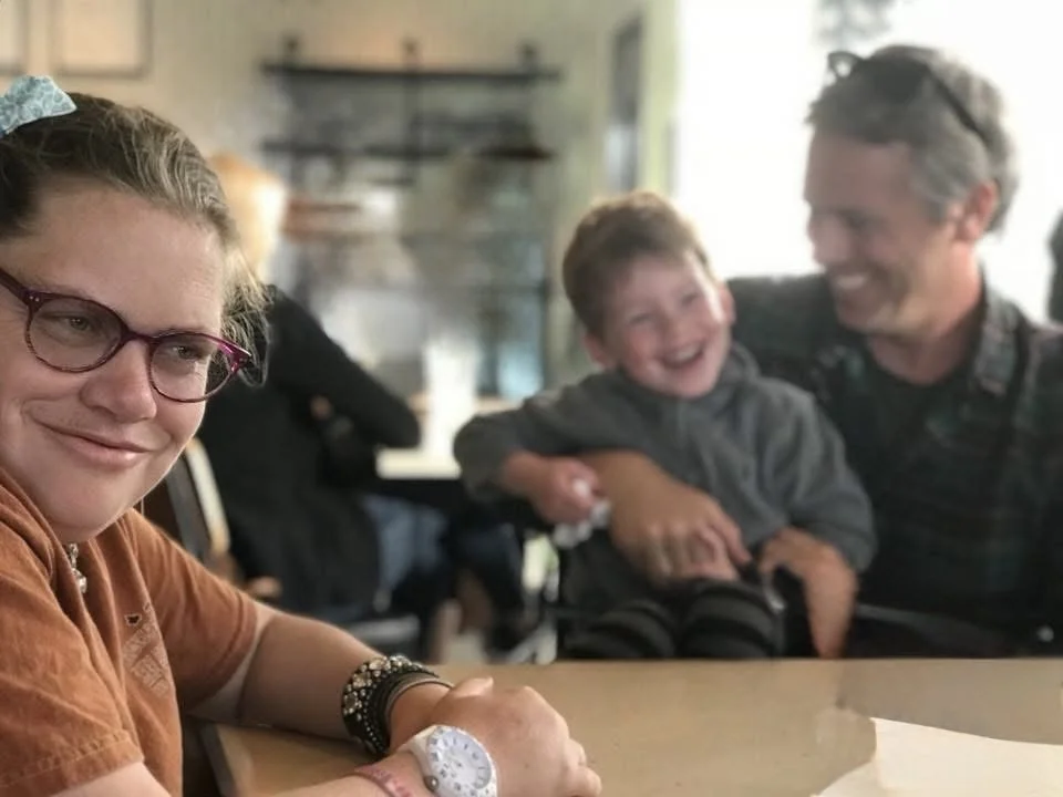 A group of four people, two adults and two children, smiling and laughing in a casual indoor setting.