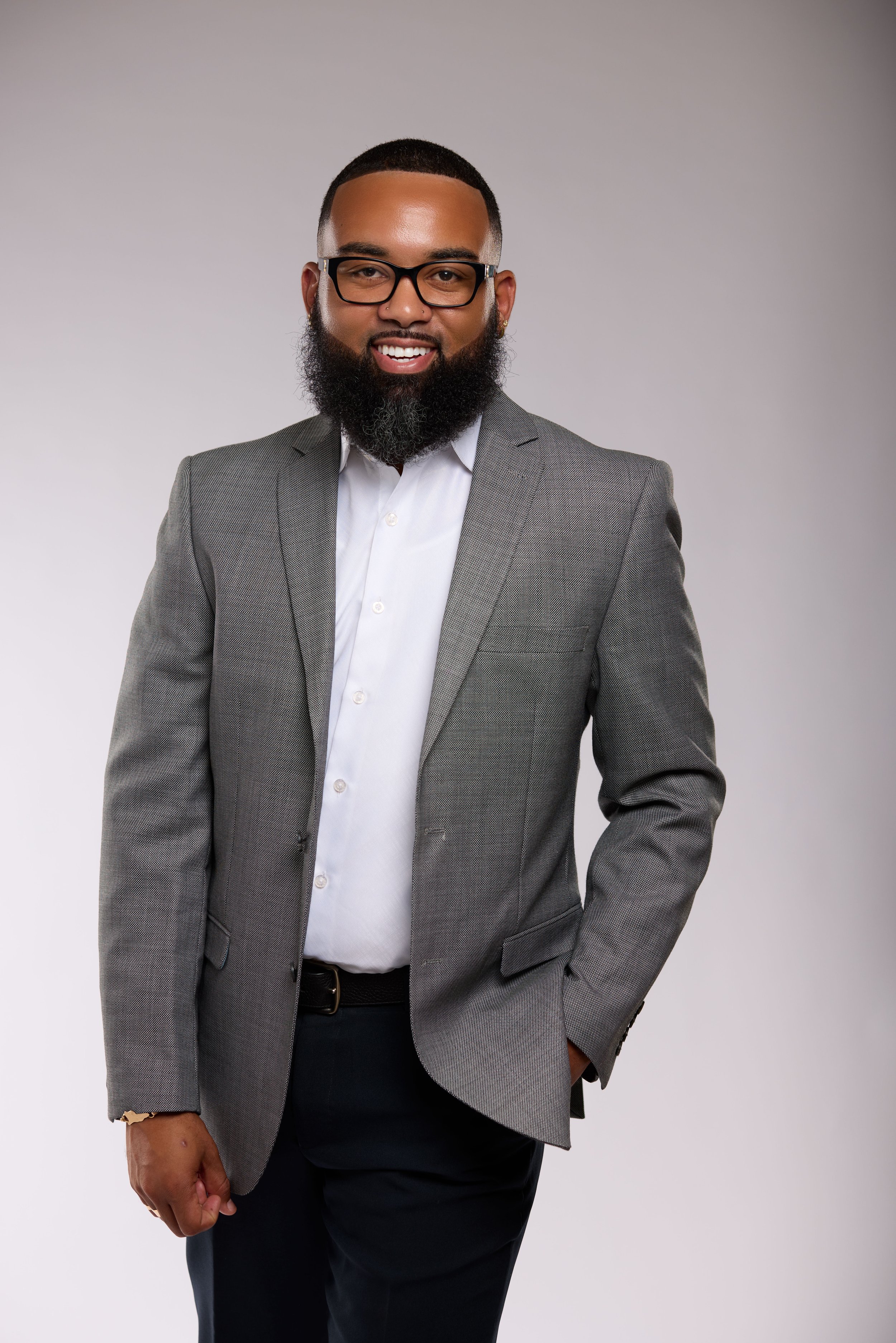 Portrait of a smiling man with glasses, a beard, wearing a gray blazer and white shirt, standing against a plain background.