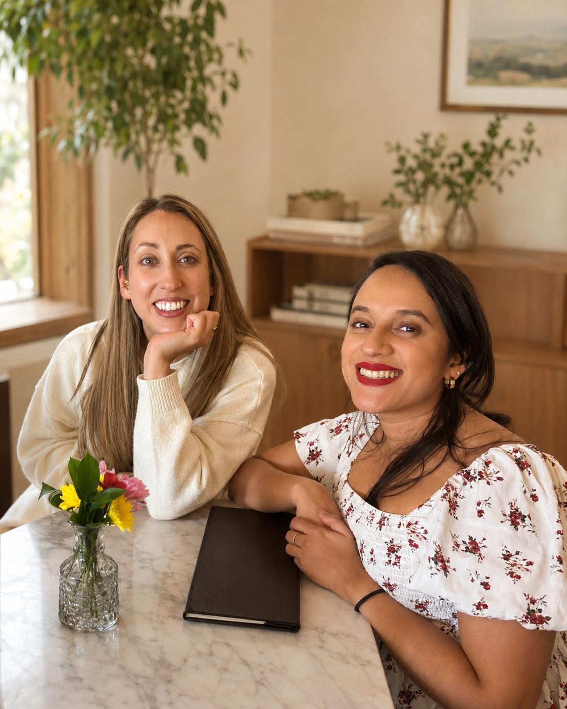 Two women sitting at a marble table with a small vase of flowers, smiling, in a cozy, well-lit room with wooden furniture and green plants.