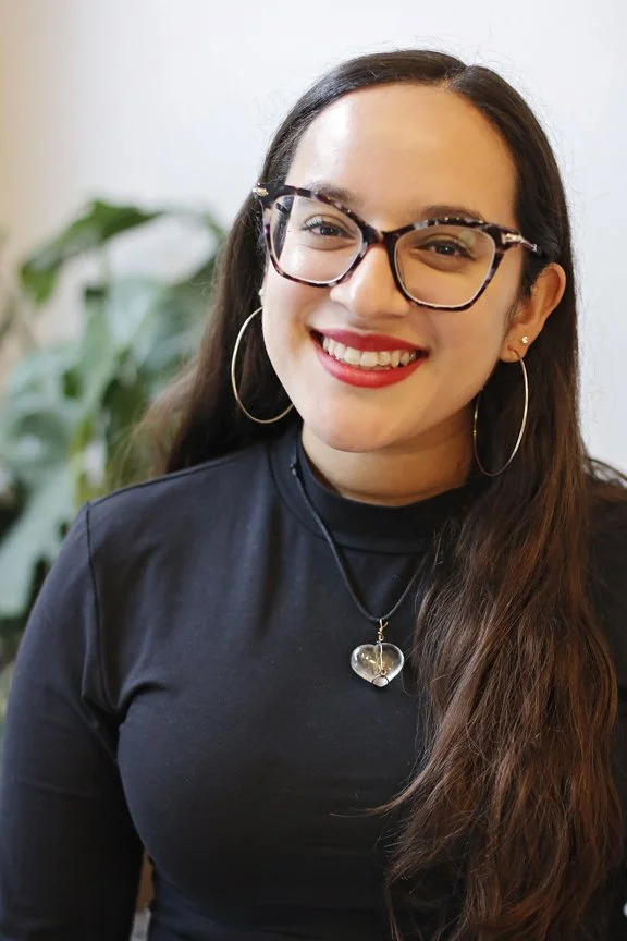 A smiling woman with long dark hair wearing glasses, hoop earrings, a black top, and a heart-shaped pendant necklace, standing in front of a blurred green plant background.