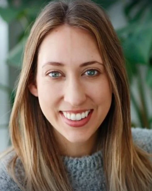 Close-up of a smiling young woman with long light brown hair and blue eyes, wearing a gray sweater, with green plants in the background.