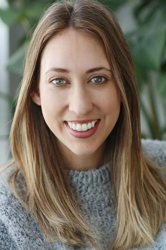 A smiling young woman with light brown hair and blue eyes, wearing a gray knitted sweater, standing near green plants.