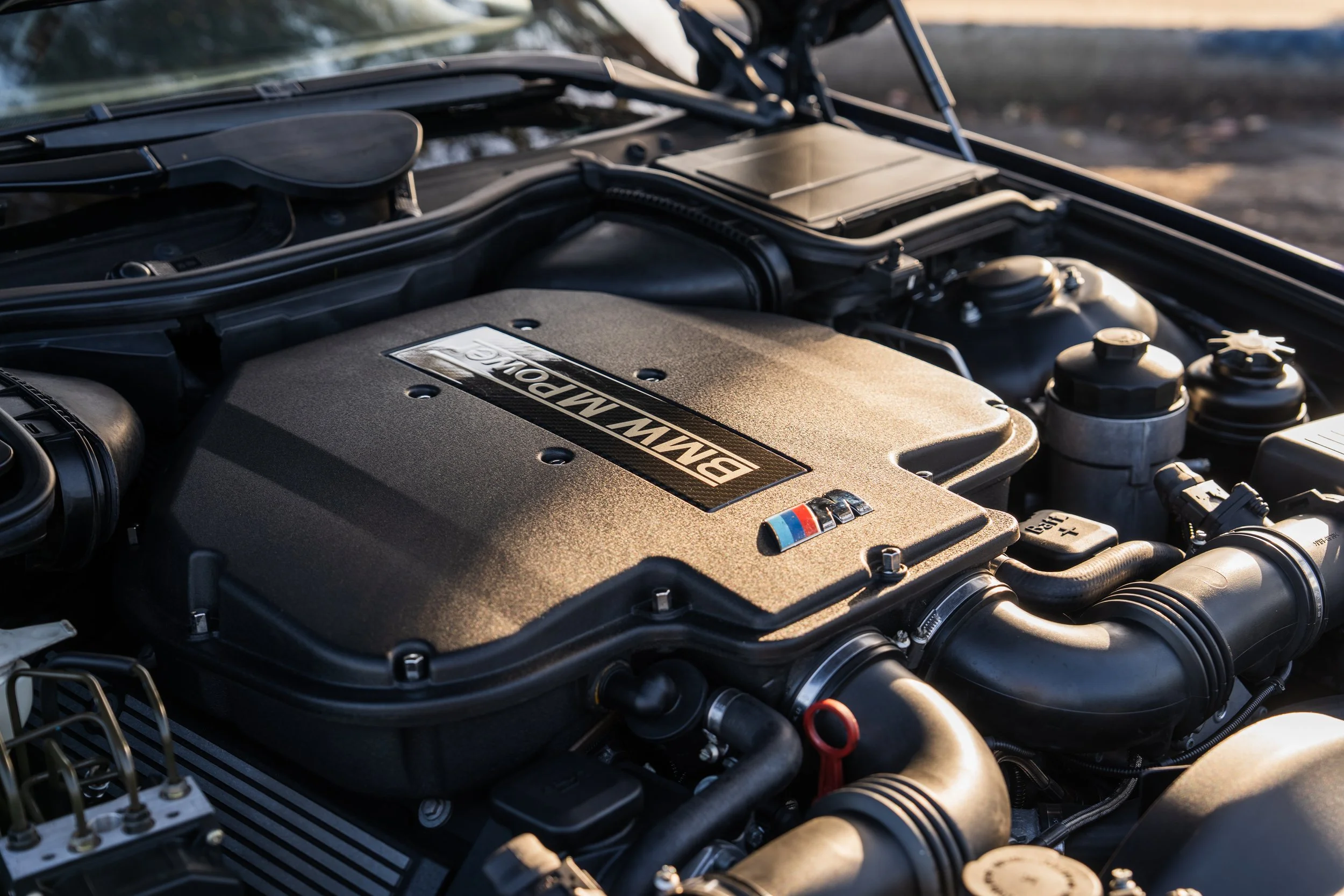 Close-up of an E39-generation BMW M5 engine with a black plastic cover displaying the BMW logo and M series badge, various engine components and hoses visible.