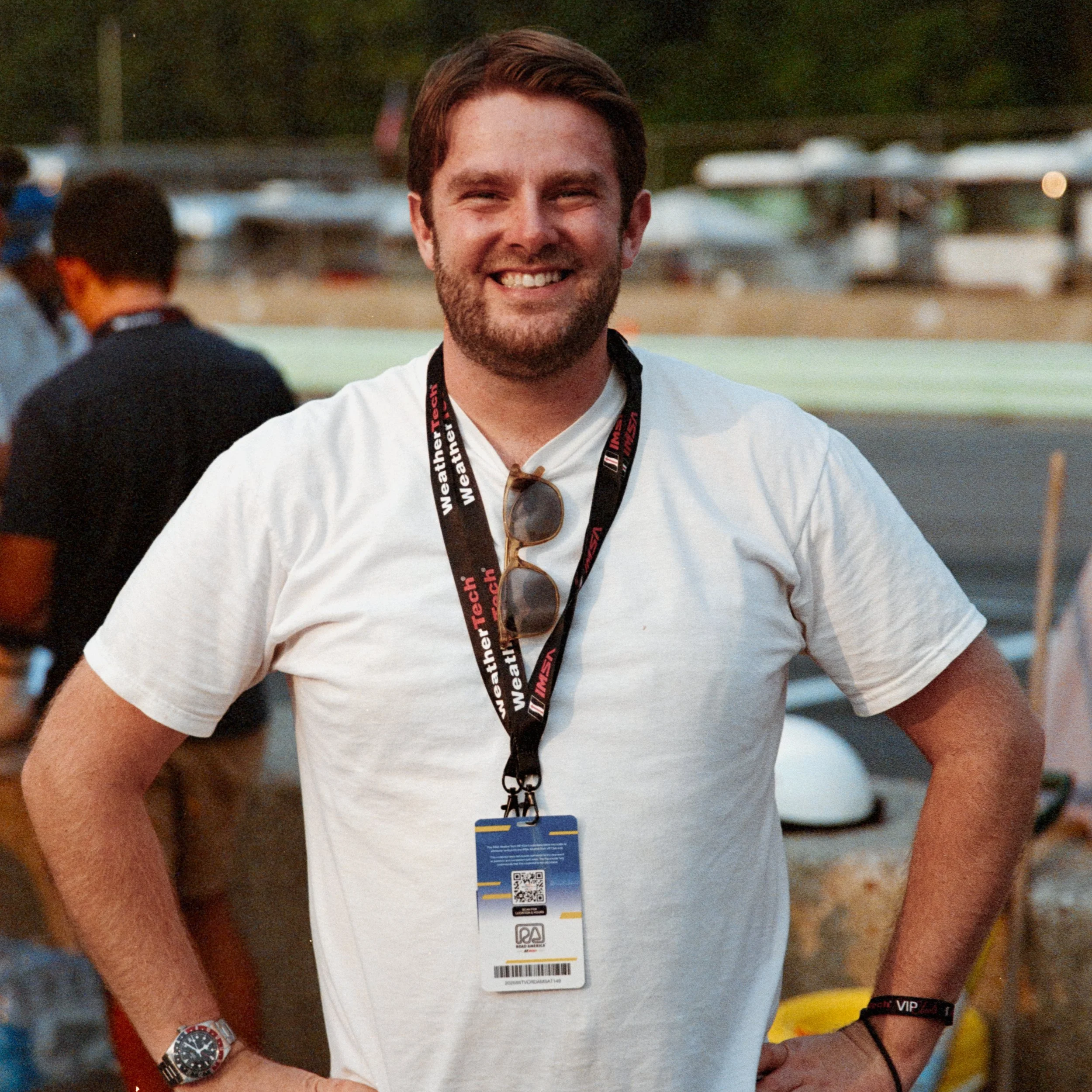 A smiling man wearing a white shirt, sunglasses hanging on his collar, a wristwatch, and a black wristband, standing outdoors with blurred people and a parking lot in the background.