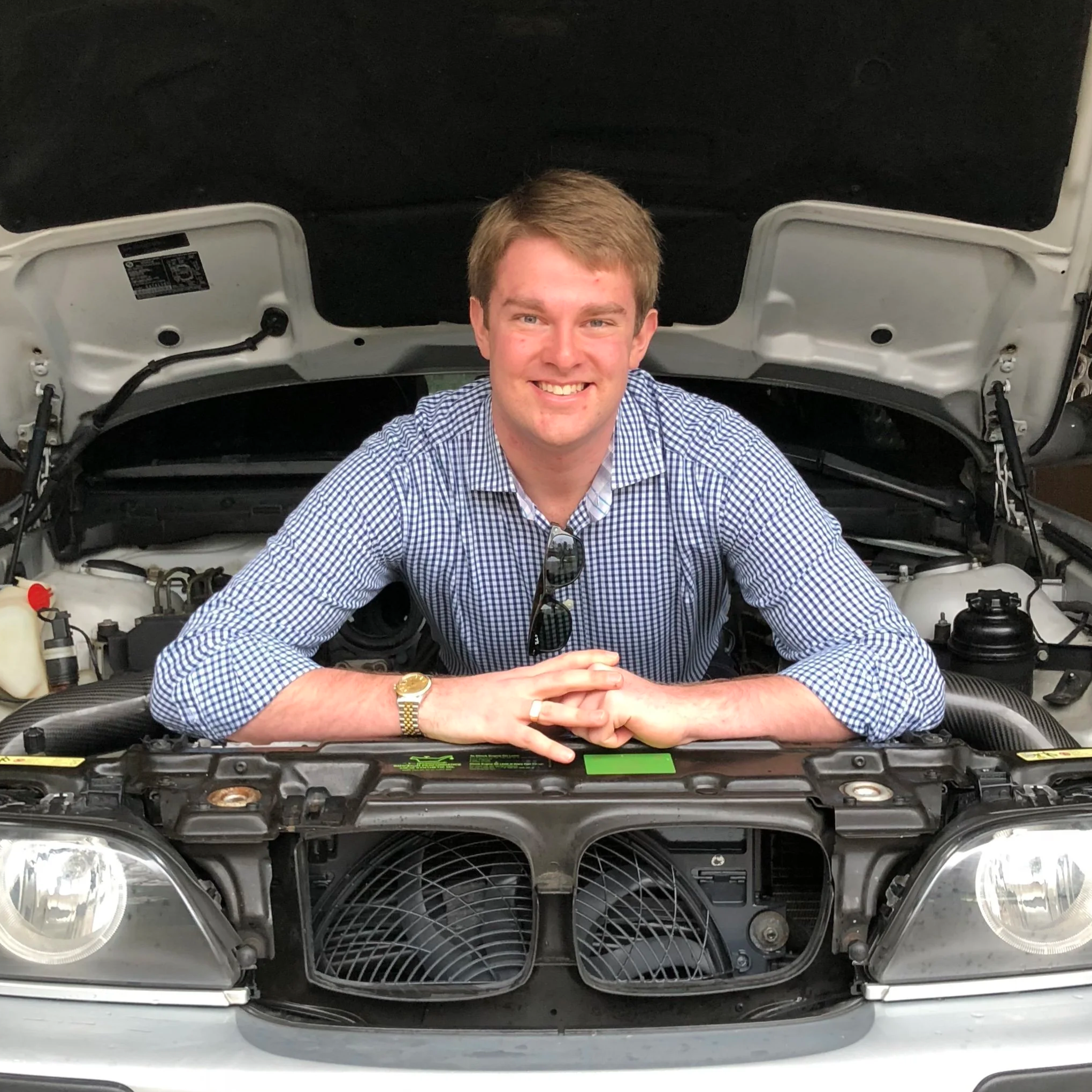 Young man smiling in front of an E39 BMW M5 with the hood open, showing engine components.