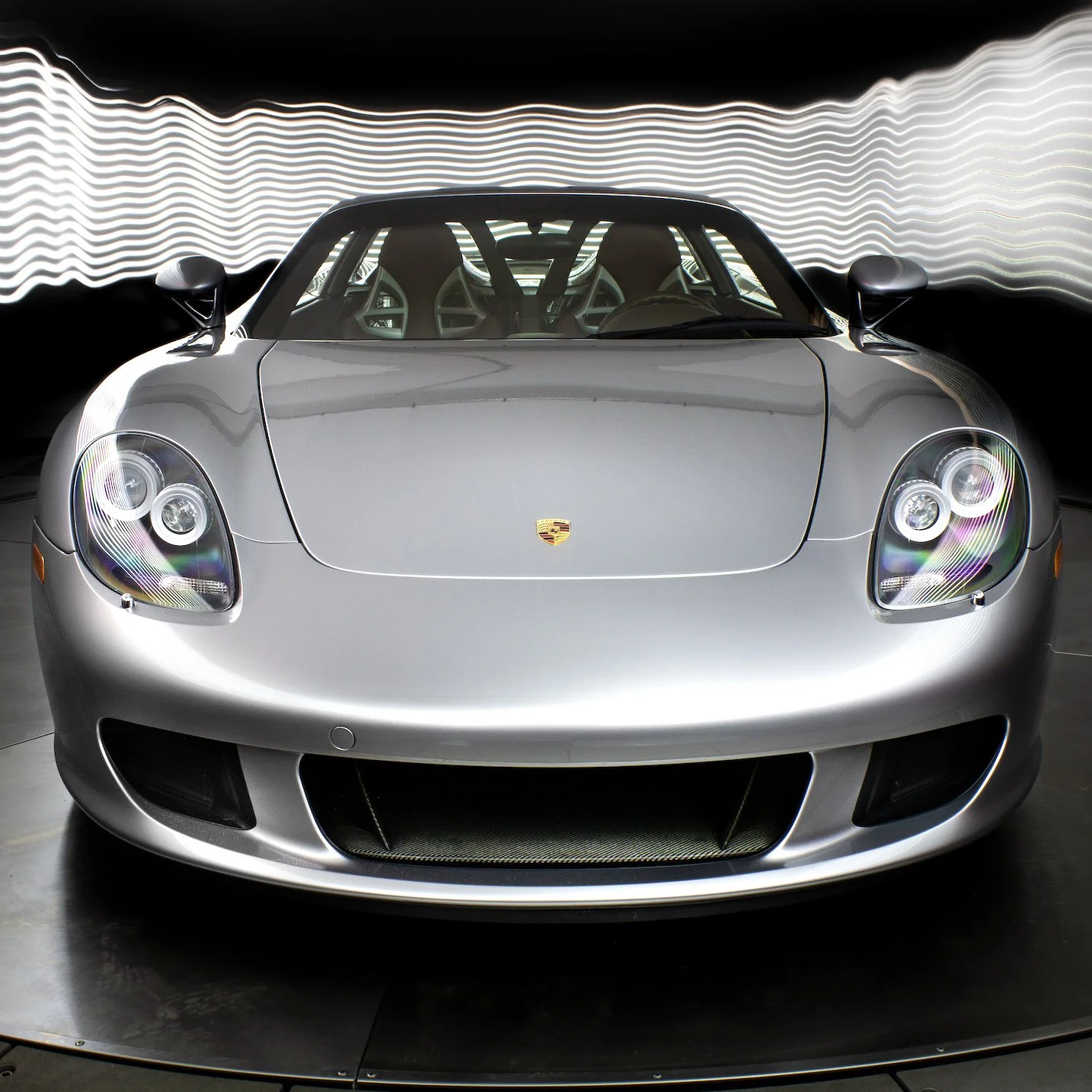 Front view of a GT Silver Porsche Carrera GT with black side mirrors and a black roof, parked indoors with a wavy light pattern background.