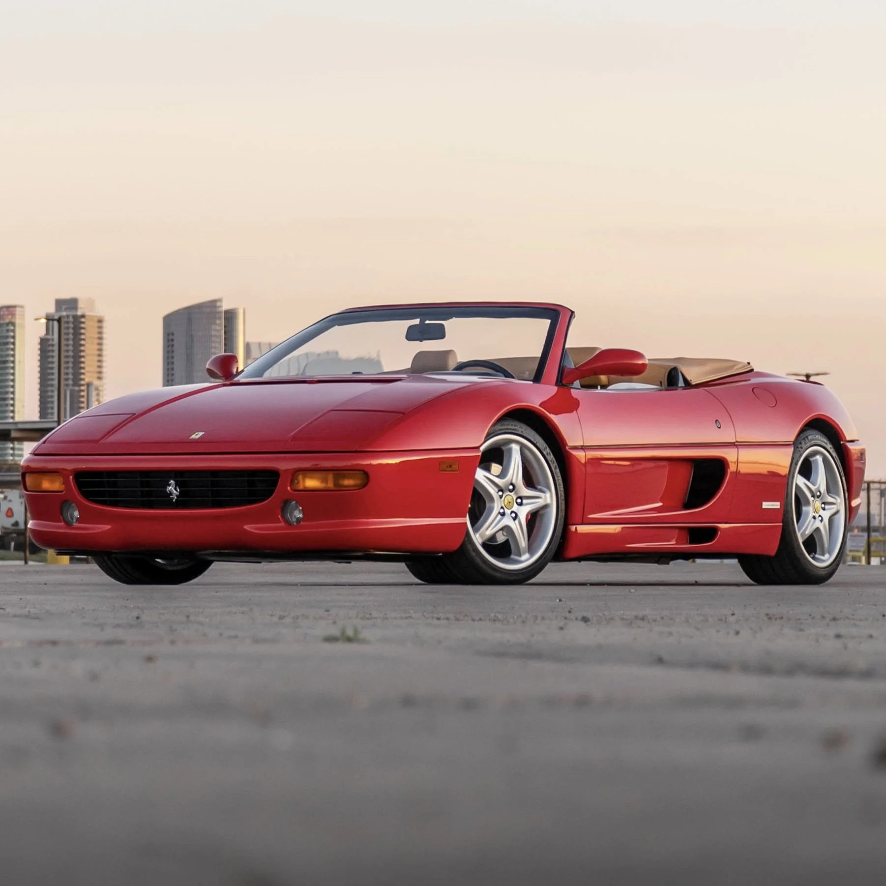 A red Ferrari 355 F1 Spider parked on a flat surface with a cityscape of San Diego in the background during sunset or sunrise.