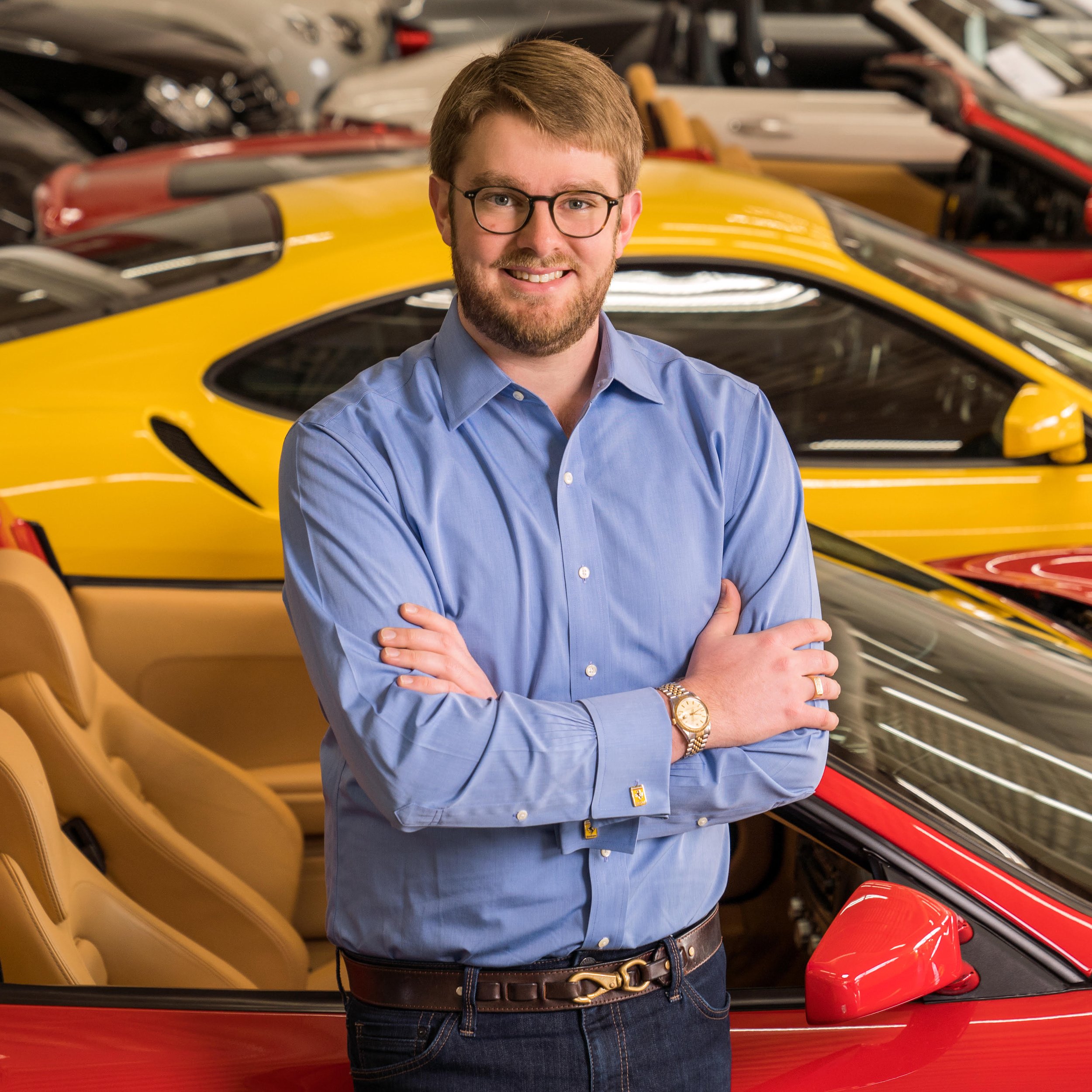 A man with glasses, a beard, and wearing a blue shirt, stands with his arms crossed in front of a row of sports cars in a showroom or dealership.