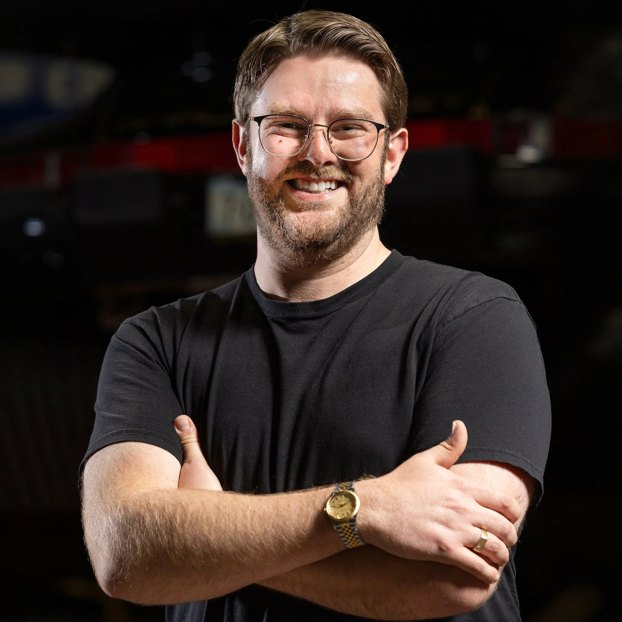 A man with brown hair, glasses, and a beard smiling, crossing his arms, wearing a black t-shirt and a Rolex Oyster Date on his right wrist, standing against a dark background.