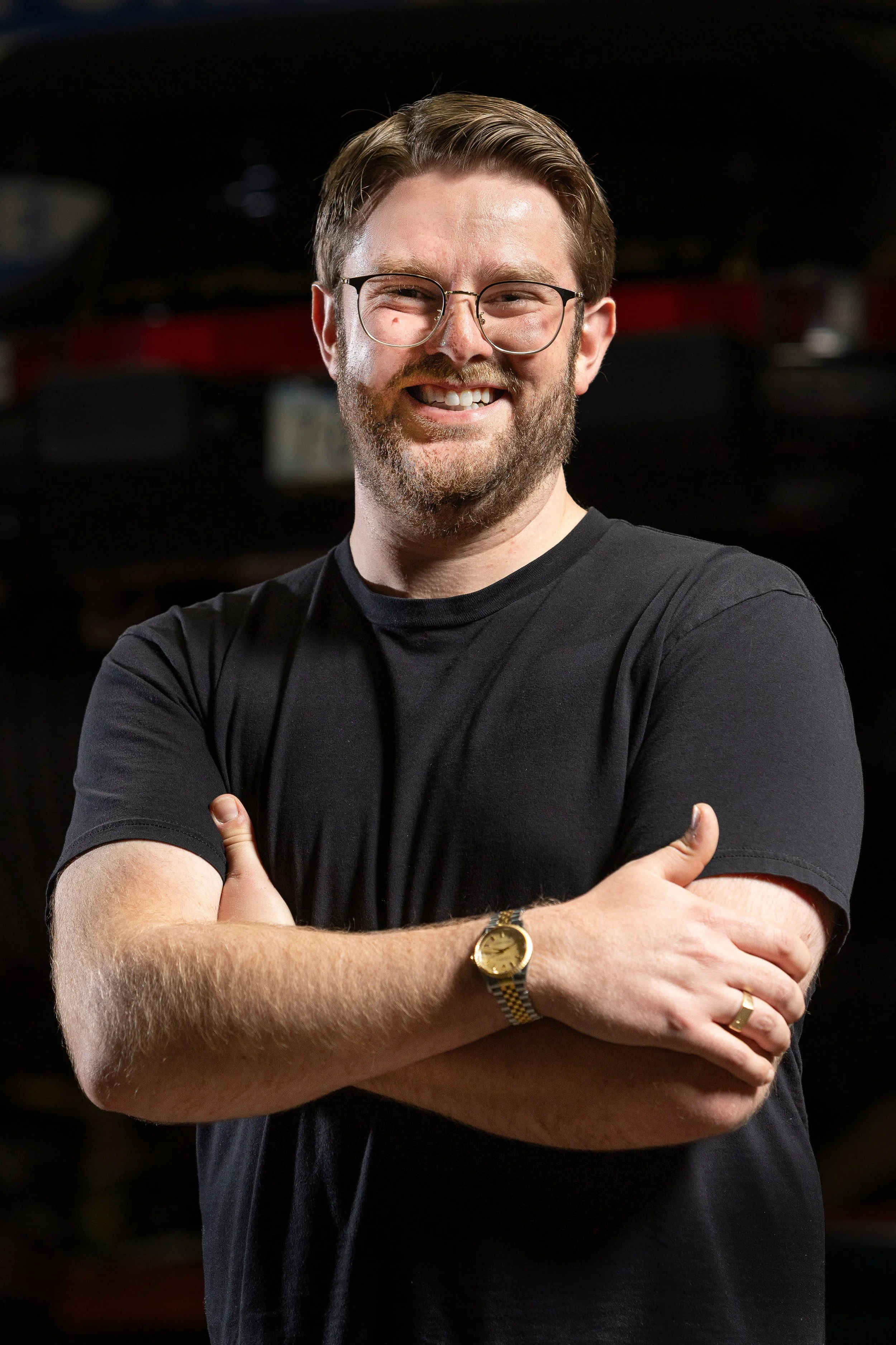 A young man with glasses, a beard, and short brown hair smiling, crossing his arms, wearing a black T-shirt and a Rolex Oyster Date, standing in front of a dark background.