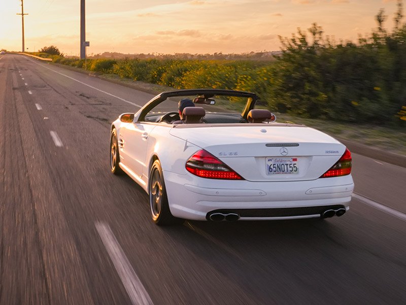 White Mercedes-Benz convertible driving on a two-lane road during sunset, with green bushes and yellow flowers on the roadside.