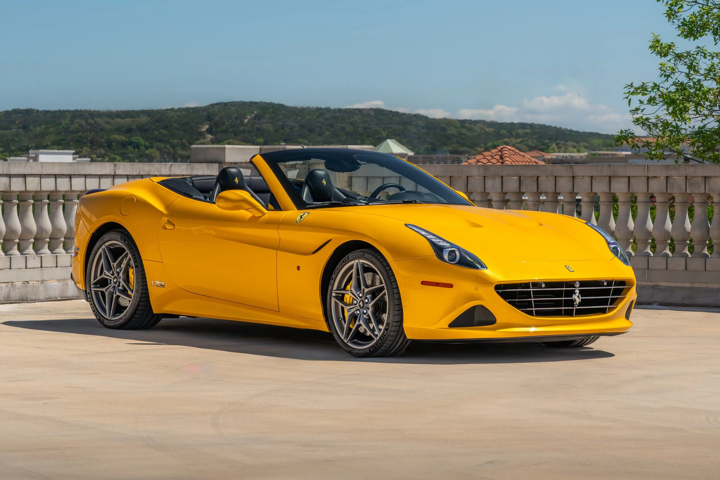 A Giallo Triplo Strada Ferrari California T 70th Anniversary parked on a rooftop with a scenic view of hills and buildings in the background.