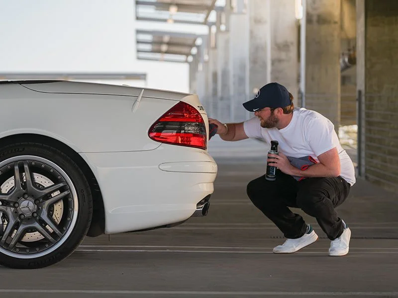 A man in a white t-shirt, black pants, white sneakers, and a dark baseball cap is crouching down to clean the tail lights of a Mercedes SL65 before getting professional car photographs for auctions