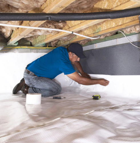 A man kneeling on a sheet of vapor barrier or plastic sheeting, installing or inspecting insulation in an attic or crawl space with exposed wooden beams and pipes overhead.