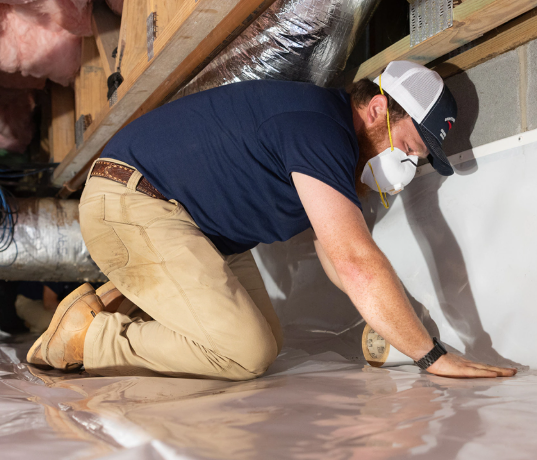 Man kneeling on the floor in a building, wearing a mask and a cap, working on the floor with tools.