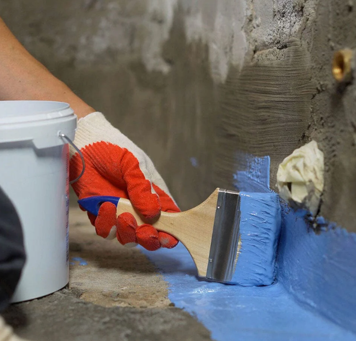 Person applying blue paint to a concrete wall using a paintbrush while wearing a red work glove.