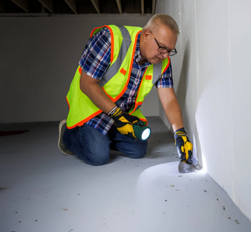 A man in safety vest and gloves kneeling and inspecting the floor with a flashlight in a building interior.
