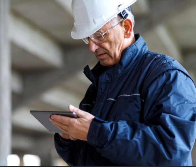 A construction worker wearing a white safety helmet and glasses, looking at a tablet in an unfinished building.