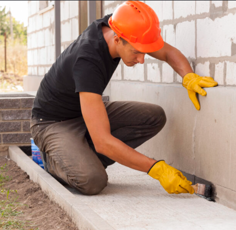 A construction worker wearing an orange safety helmet and yellow gloves is applying a coat of gray paint or stain to a concrete sidewalk beside a building under construction.