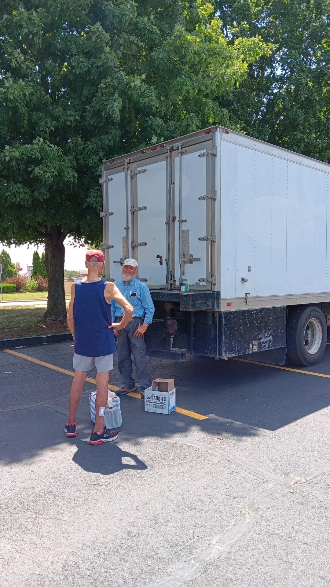 Two men are having a conversation beside a large white moving truck parked in a lot, with a tree in the background. One man is dressed in casual athletic wear, and the other is wearing a blue button-up shirt and gray pants. There are boxes and bags on the ground near them.