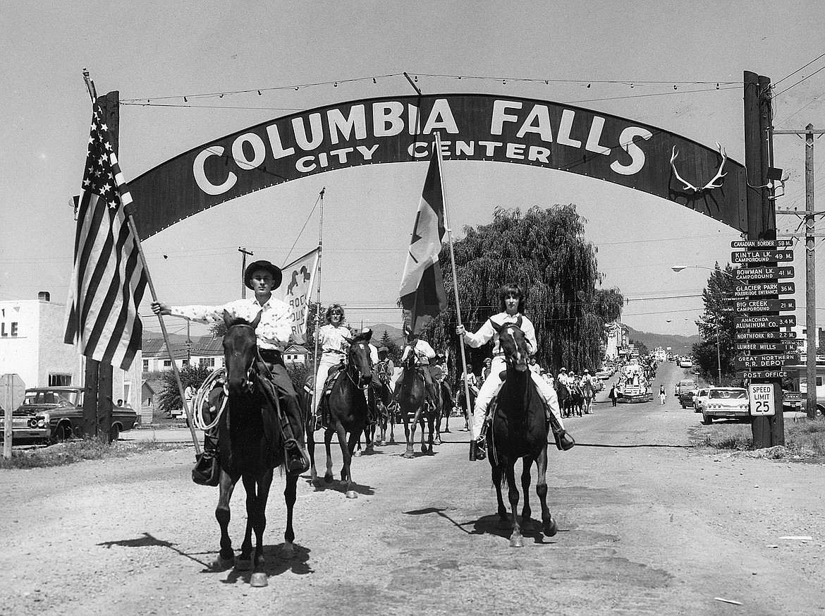 A parade scene at Columbia Falls City Center with people on horseback carrying American flags and a flag with a bear image, under an archway with the city name, surrounded by vintage cars and street signs.