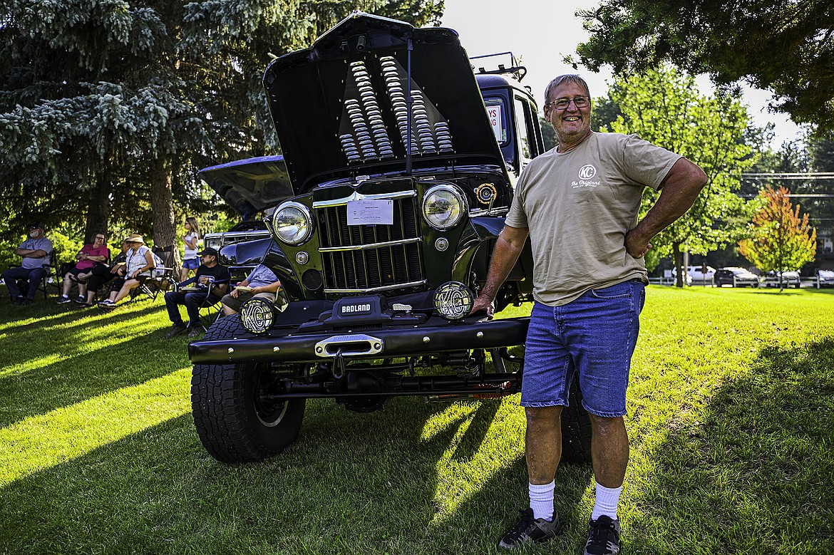 A man smiling and standing with his hand on his hip next to a black classic car with an open hood, at an outdoor event on a grassy area with trees and people sitting on chairs in the background.