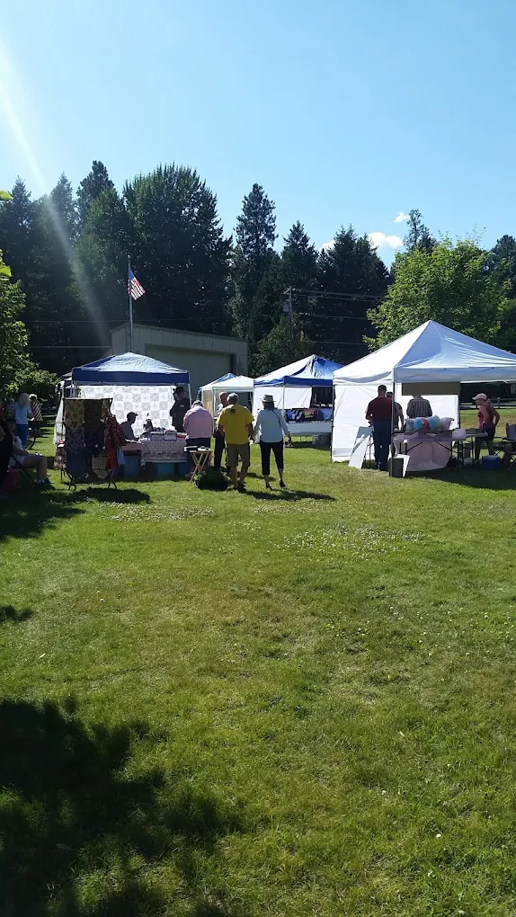 Outdoor market or fair with white tents, people walking and browsing, grassy area, trees and a building in the background, and a flag flying on a pole.