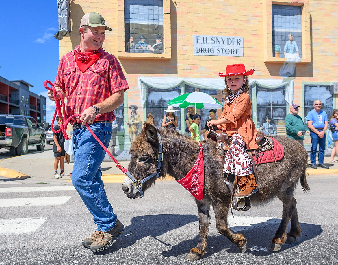A young girl riding a small donkey, being led by a man, in a parade on a sunny day in front of a brick building with a sign that reads "E.H. Snyder Drug Store." The girl wears a red cowboy hat and orange shirt, and the donkey has a red bandana around its neck.