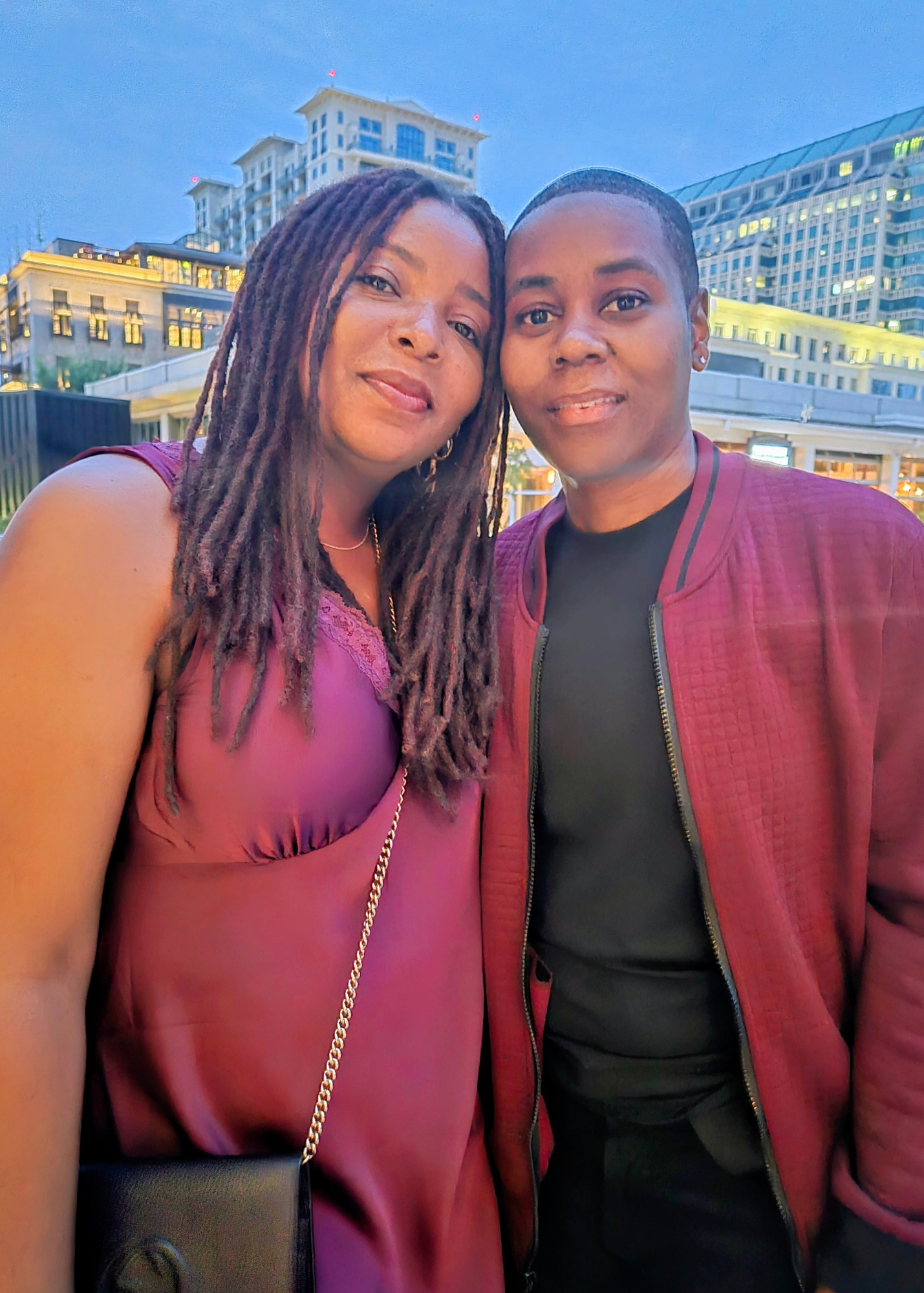 Two women taking a selfie outdoors in front of a cityscape with tall buildings and a clear evening sky.