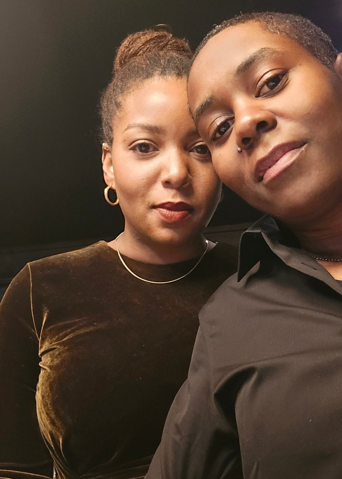 Close-up selfie of two women posing together indoors, with black background. One woman has short hair, nose piercing, and wears a black shirt. The other woman has her hair styled in a bun and wears a dark velvet top with gold hoop earrings.