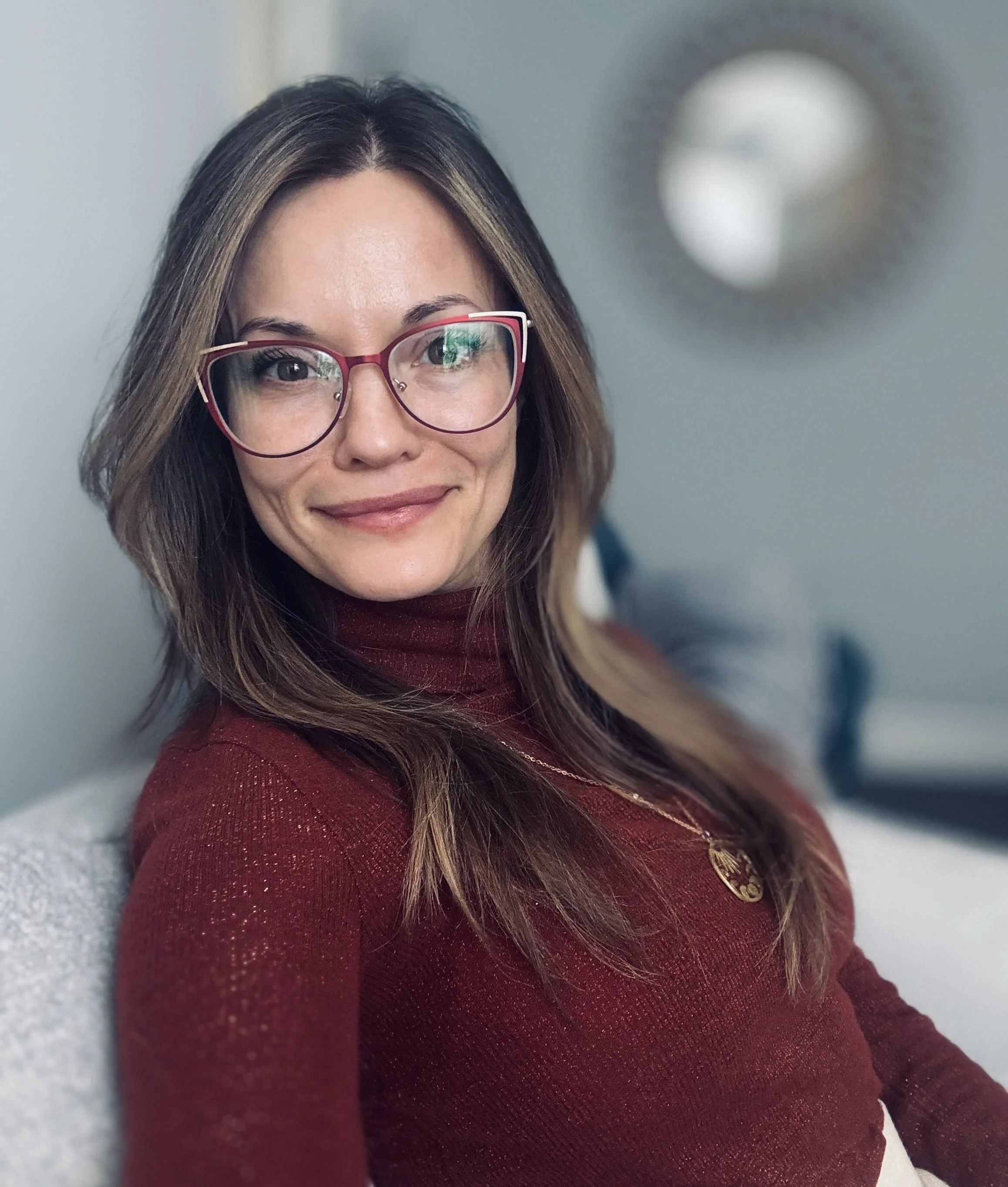 A woman with long brown hair wearing red-rimmed glasses and a maroon sweater, smiling softly in a close-up photo.