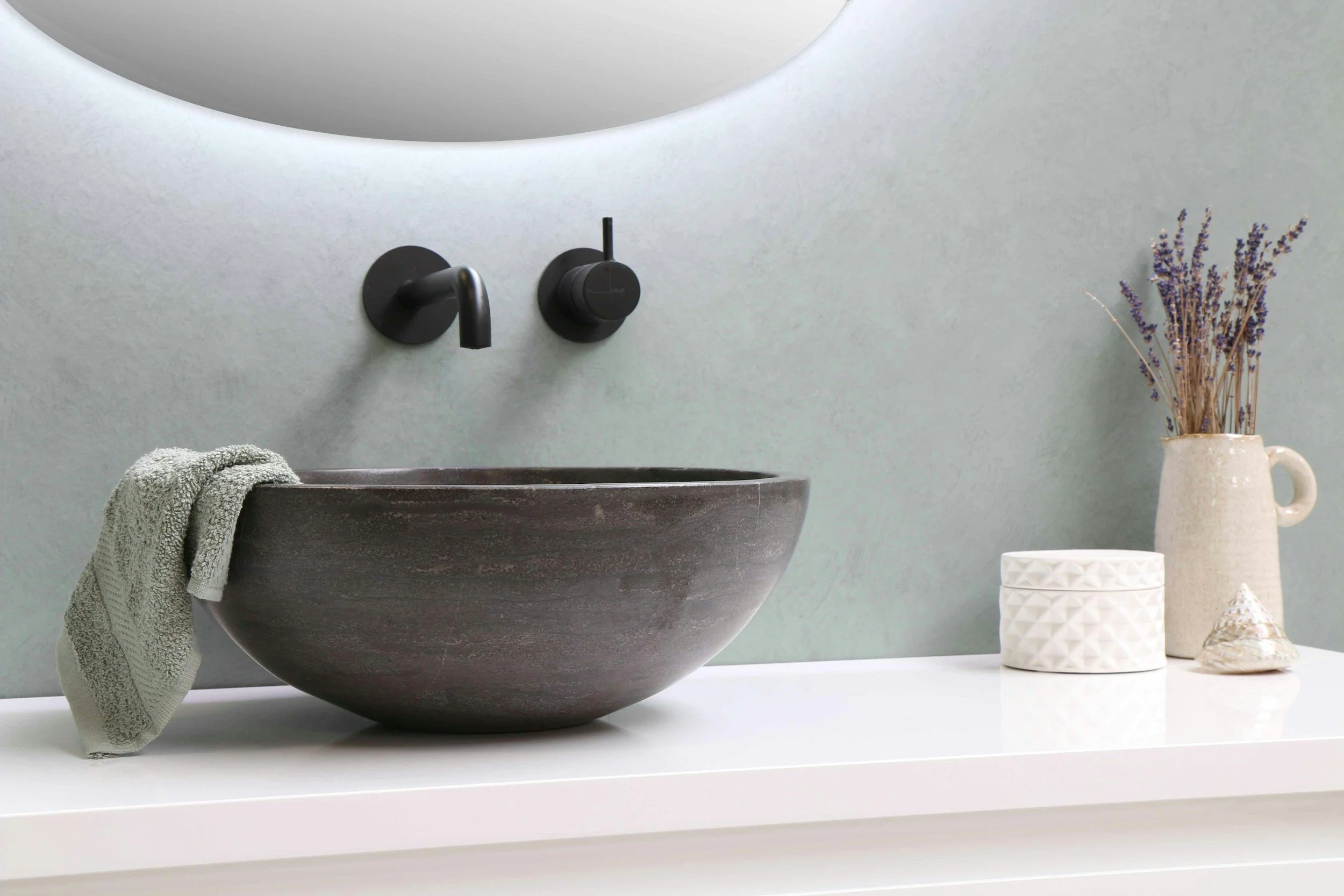 A modern bathroom sink setup with a rounded dark gray stone basin, black matte faucet, and matching black knobs, with a vintage-style white ceramic vase filled with dried lavender, a small patterned cup, and a seashell on a white countertop against a light neutral wall.