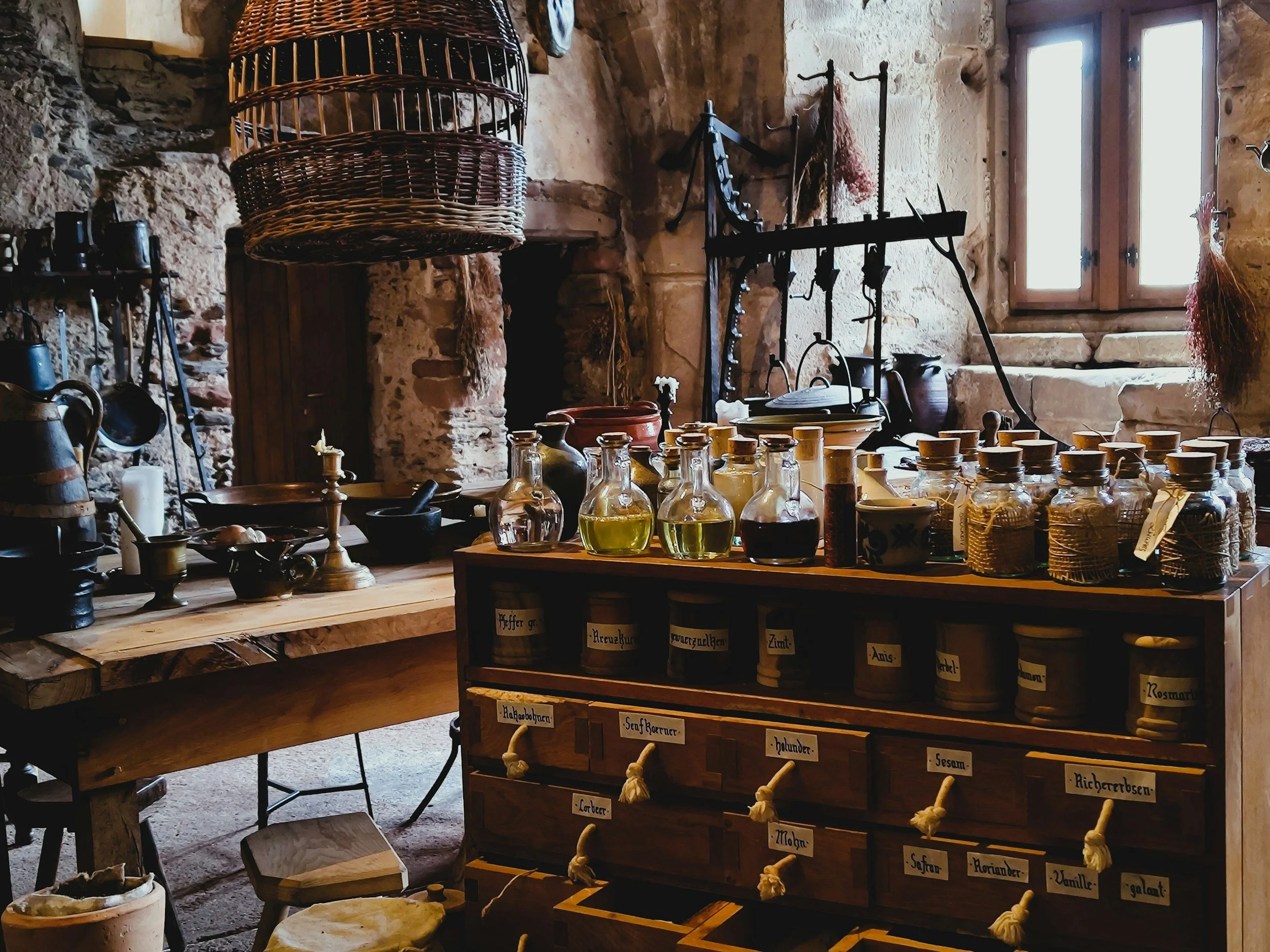 A rustic kitchen or apothecary scene with stone walls, a wooden table, glass bottles filled with liquids, a large wicker basket, and various kitchen utensils and jars with labels.