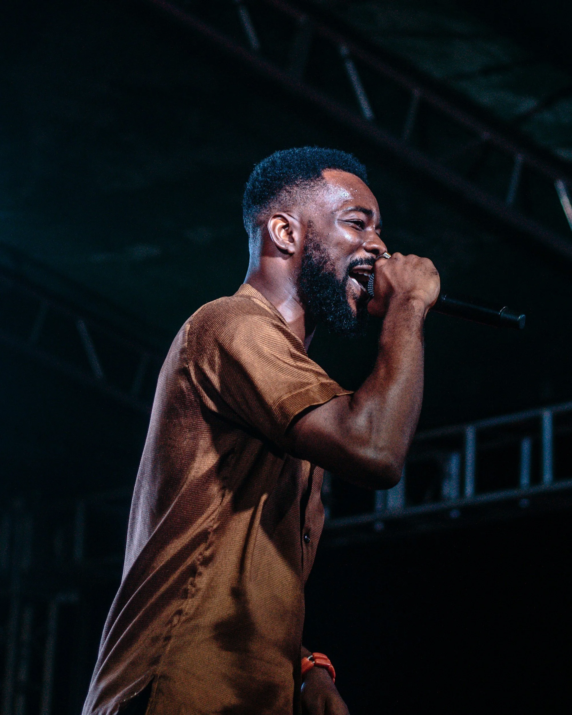 Man singing into a microphone during a performance, with dark background and stage lighting.