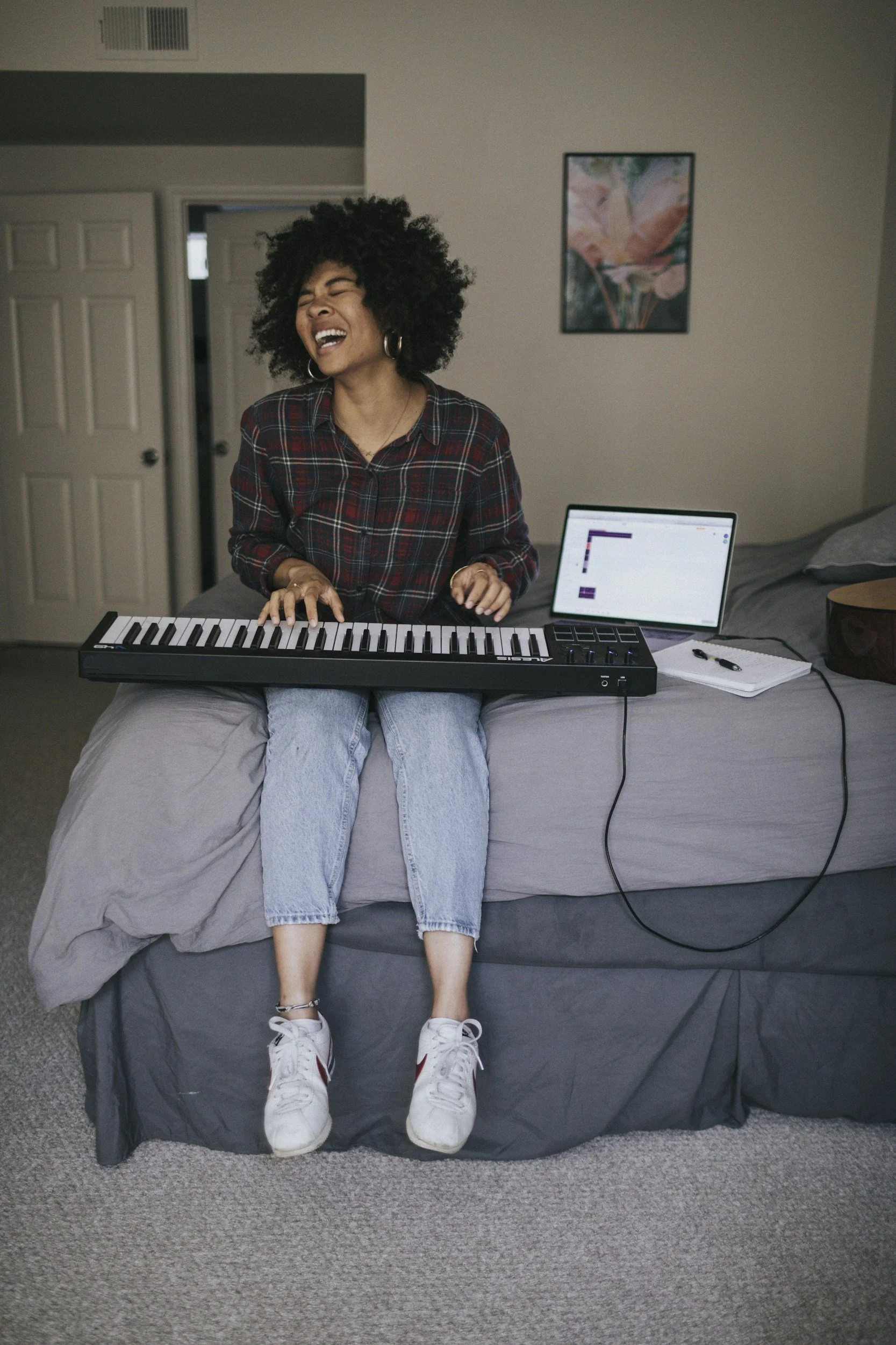 A woman with curly black hair singing while playing a keyboard in a bedroom. She is wearing a plaid shirt, light blue jeans, and white sneakers. There is a laptop, notebook, and pen on the bed beside her. The background shows a closed door, a painting on the wall, and a bed with gray bedding.