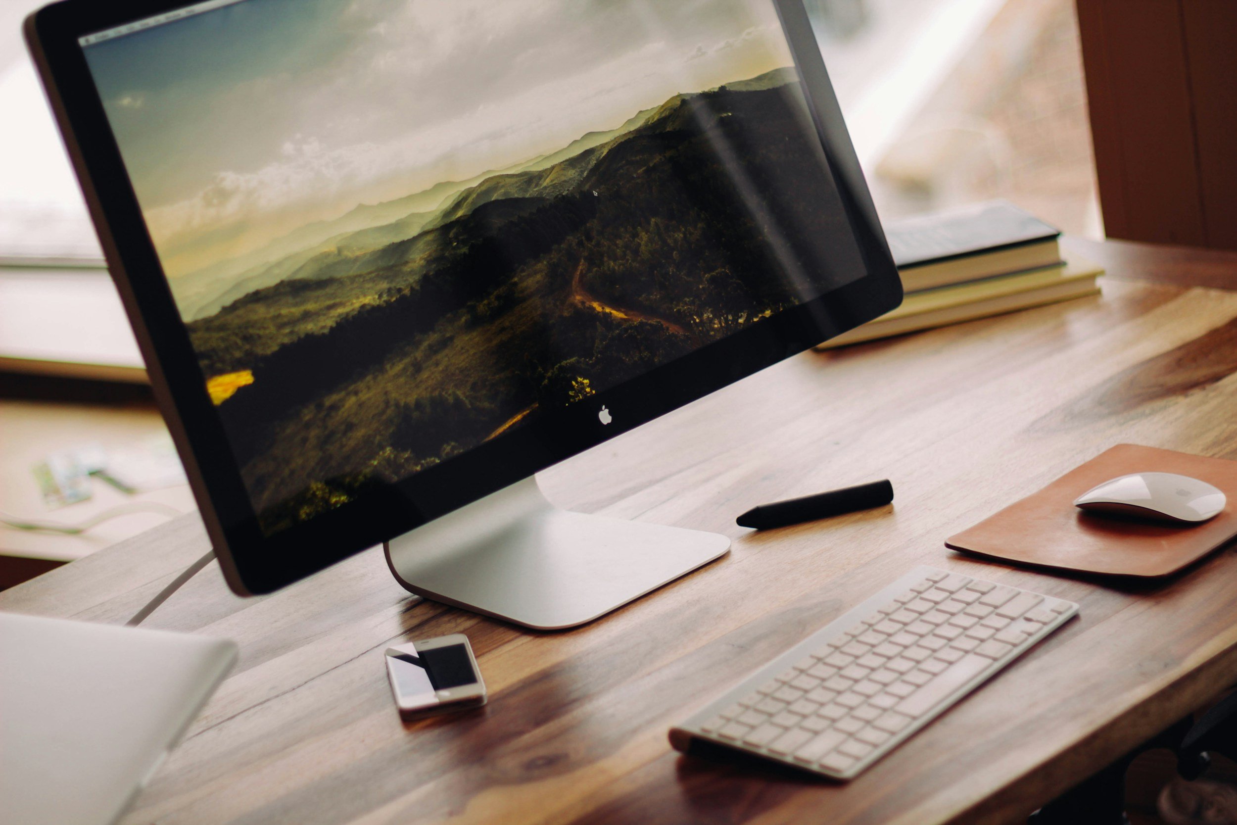 An Apple desktop computer displaying a landscape of green mountains and a cloudy sky, situated on a wooden desk with a keyboard, mouse, and a smartphone nearby.