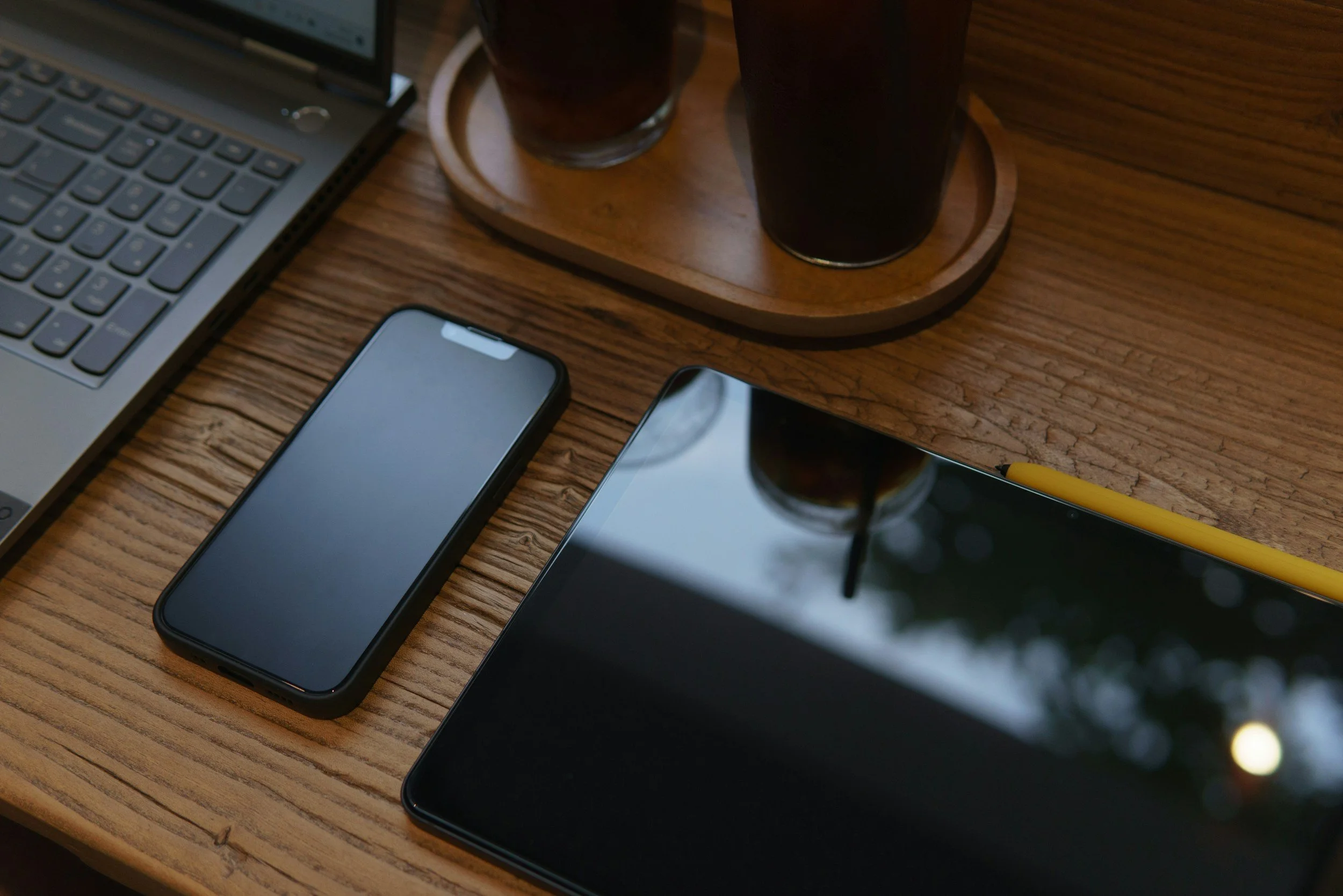 A wooden table with a laptop, an iPhone, two glasses of dark beverages on a round wooden tray, and a yellow pen.