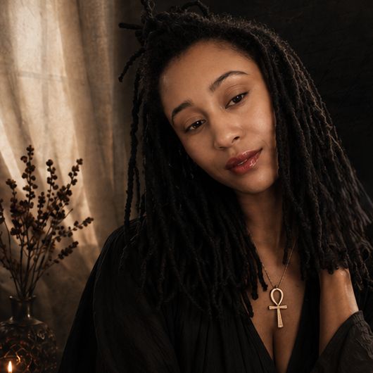 Portrait of a woman with long dreadlocks, wearing a black top and a gold ankh necklace, in a dimly lit room with a candle and plant in the background.