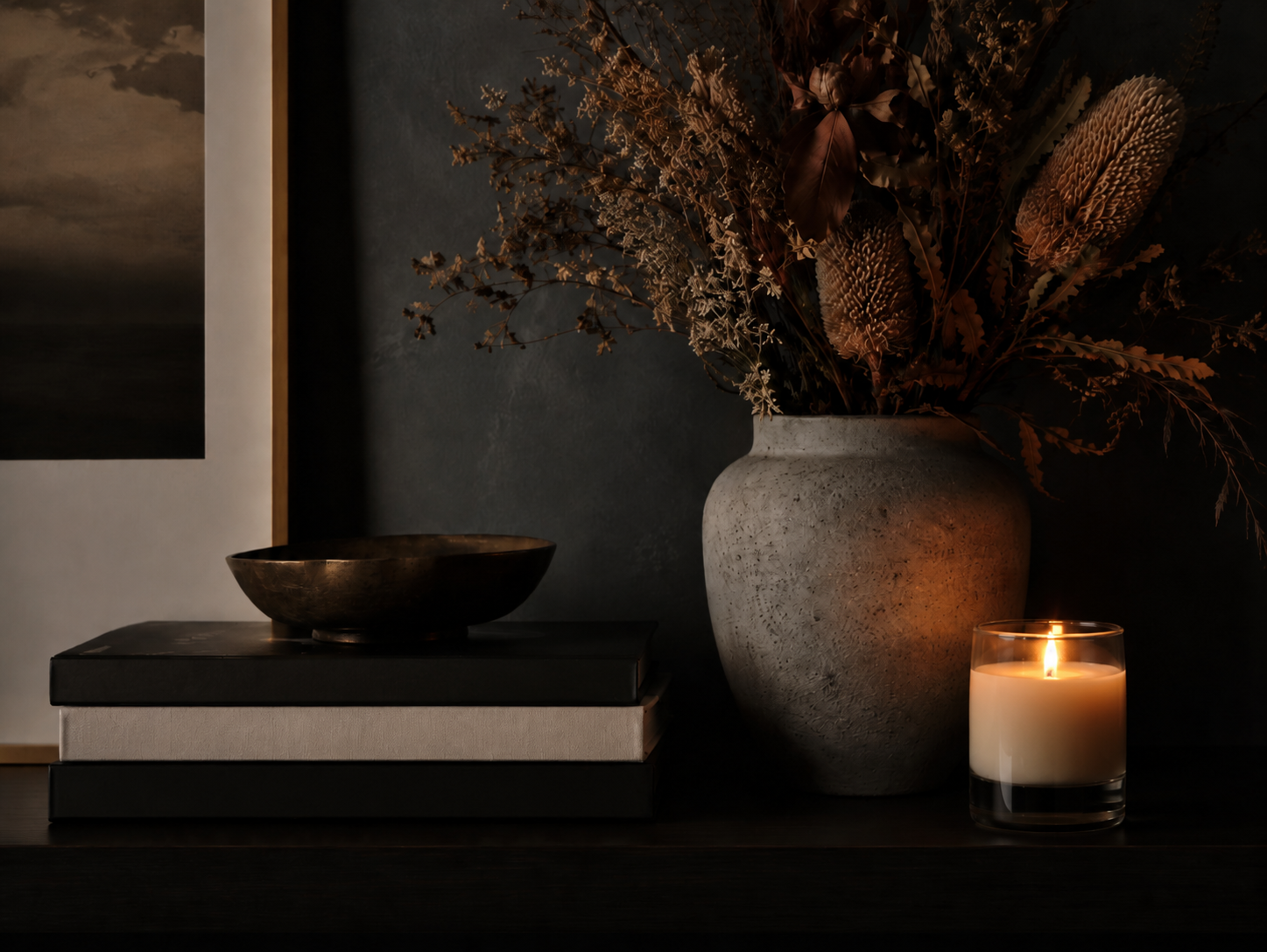 A decorated table with a large textured white vase holding dried flowers, a lit candle in a glass holder, and a couple of stacked books with a decorative bowl on top; dark background with artwork hanging on the wall.